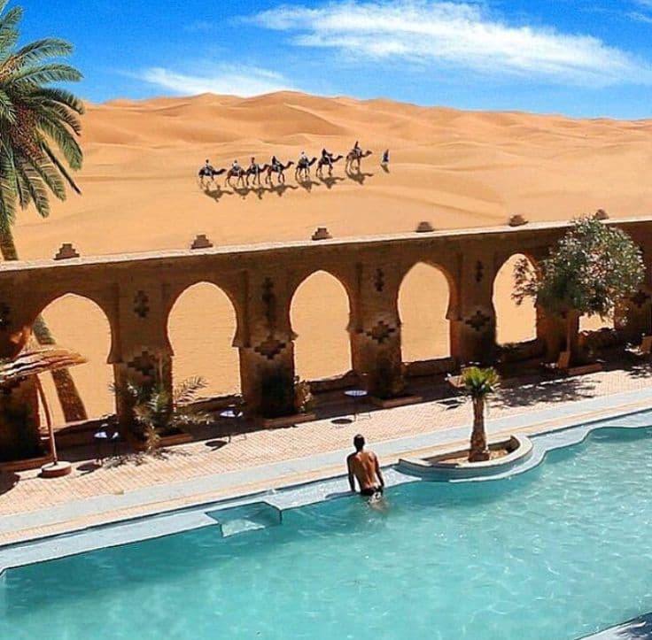 Camel caravan crossing Merzouga Sahara dunes viewed from traditional riad pool terrace in Morocco