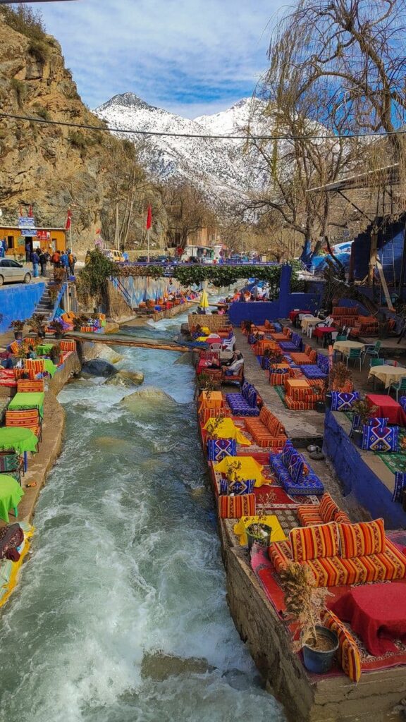 Colorful riverside cafés in Ourika Valley with snow-capped Atlas Mountains, popular day trip from Marrakech.
