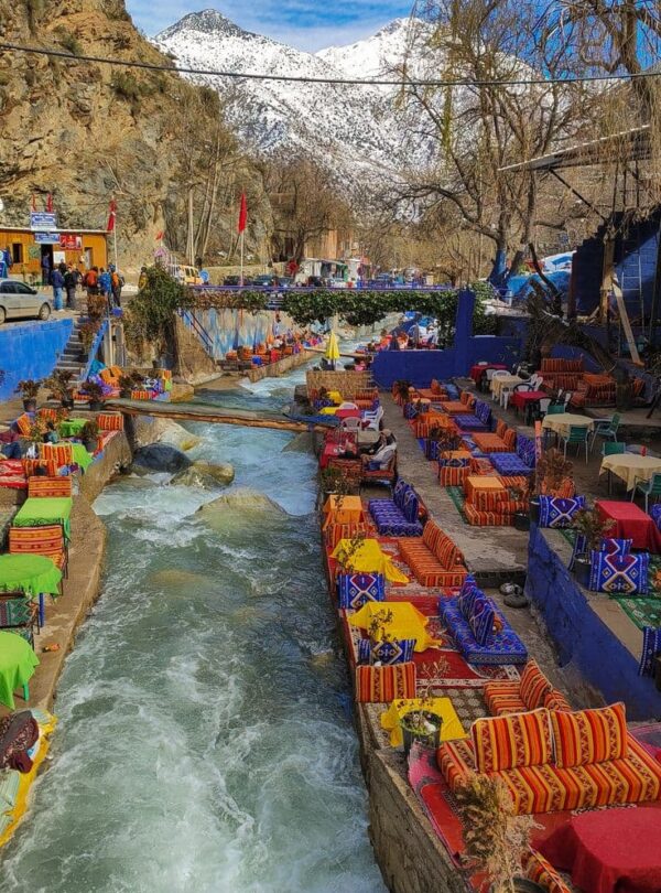 Colorful riverside cafés in Ourika Valley with snow-capped Atlas Mountains, popular day trip from Marrakech.